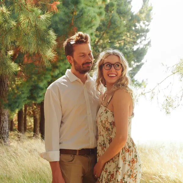 A couple standing in front of a grassy scene of pine trees. A golden shining sun on the right. Both are very happy and in love.