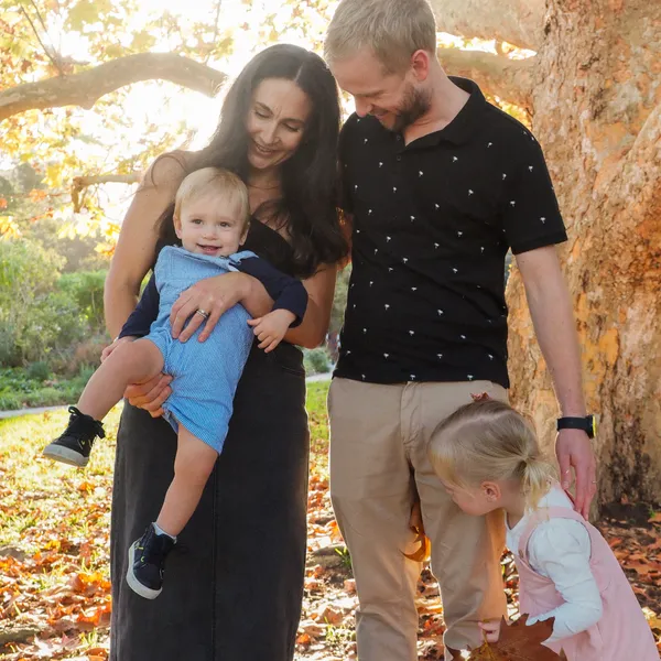 Family of 4 standing in the sun infront of an autumn tree. The younger boy is smiling and having fun being cheeky.
