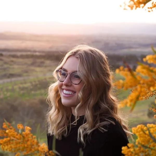 A lovely portrait of a beautiful blonde woman wearing glasses. She's wearing a black top infront of a sunny and forested background.